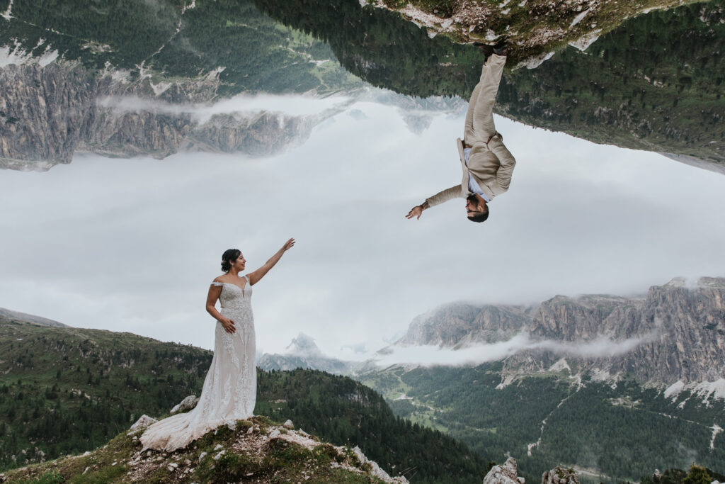 A couple reaches toward each other in an image where they are flipped upside down in the frame. They are wearing white wedding clothes with the misty, wildflower covered mountain peaks of the Dolomites in July surrounding them.