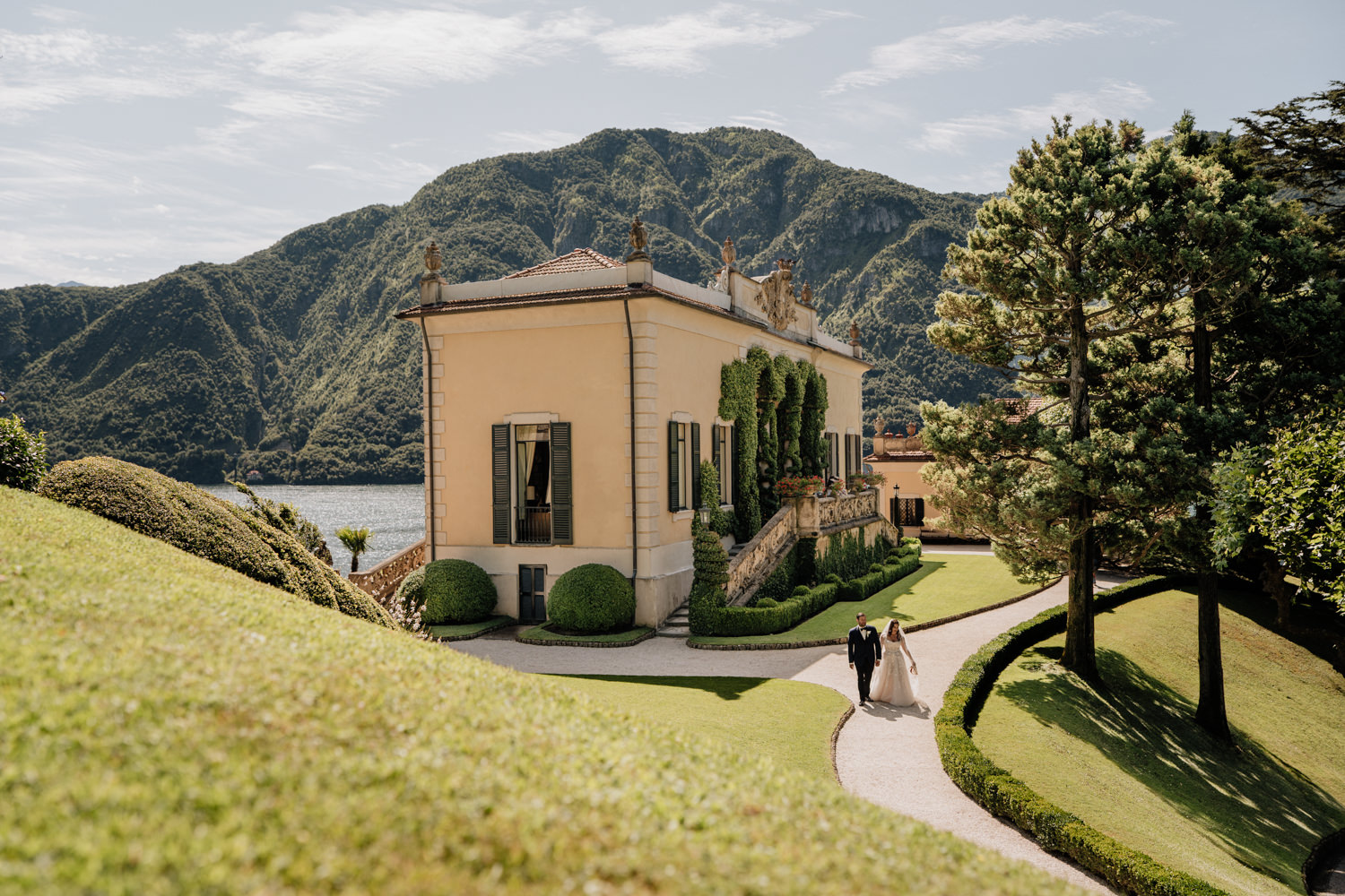 A couple in white wedding dress and black tuxedo walk in front of Villa del Balbianello during their wedding photography session on Lake Como