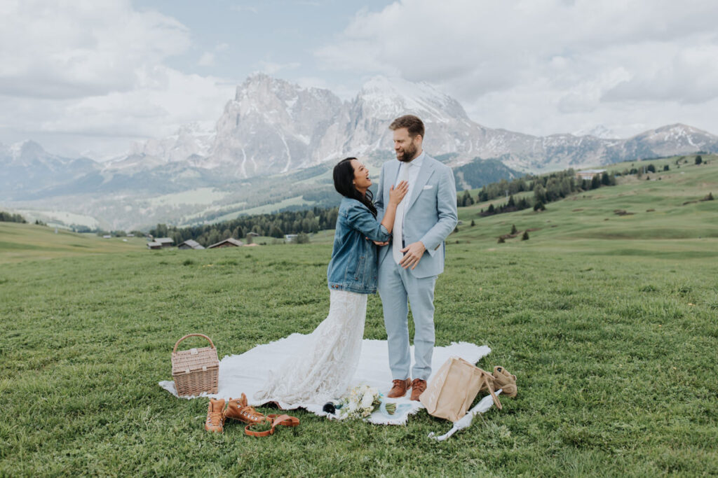 A couple pose for their elopement photos at Alpe di Suisi in the Italian Dolomites. They are standing smiling at each other on a grassy picnic setup, with snow capped mountains far in the distance.