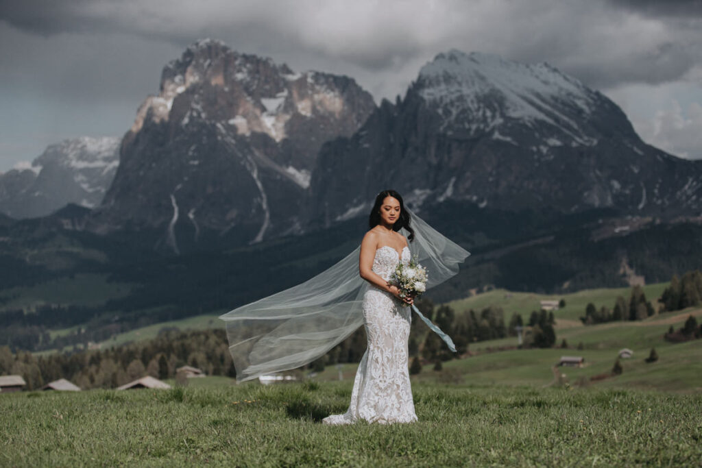 A bride in white dress and blowing wedding veil stands on the green, rolling fields of Alpe di Suisi in the Italian Dolomites for her elopement day