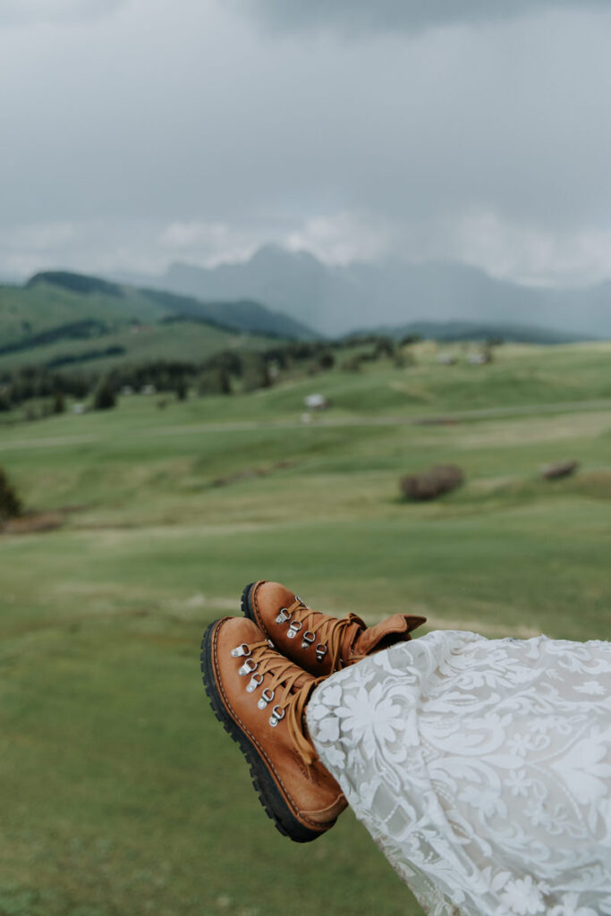 A woman's hiking boots are seen with the bottom of her wedding dress. In the background, the vibrant green meadow of Alpe di Suisi in the Italian Dolomites stretches to the mountains and a moody sky.