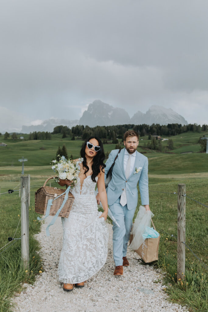 A couple walk toward the camera down a grassy trail on their elopement day at Alpe di Suisi in the Italian Dolomites. They are carrying a picnic basket, bouquet and wearing wedding clothes.