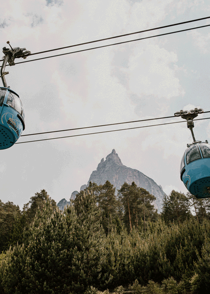 Two gondolas at the Alpe di Suisi station move past each other over tall pine trees, framing the famous Schlern peak in the background.