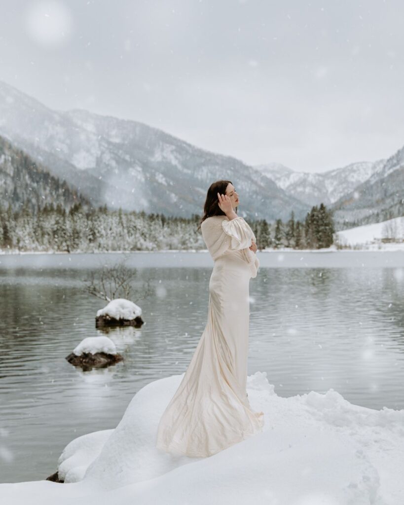 A bride in cream color wedding gown stands next to a snowy lake in Berchtesgaden, Germany on her elopement day