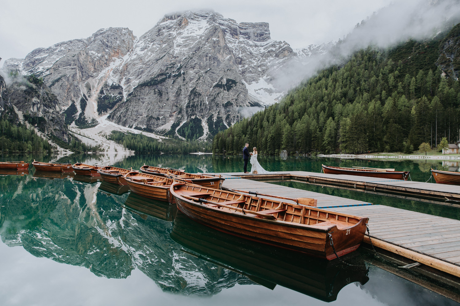 A couple read vows at the Lago di Braies boathouse on their elopement day. There is a row of wooden boats and mirrored mountain reflection surrounding them.