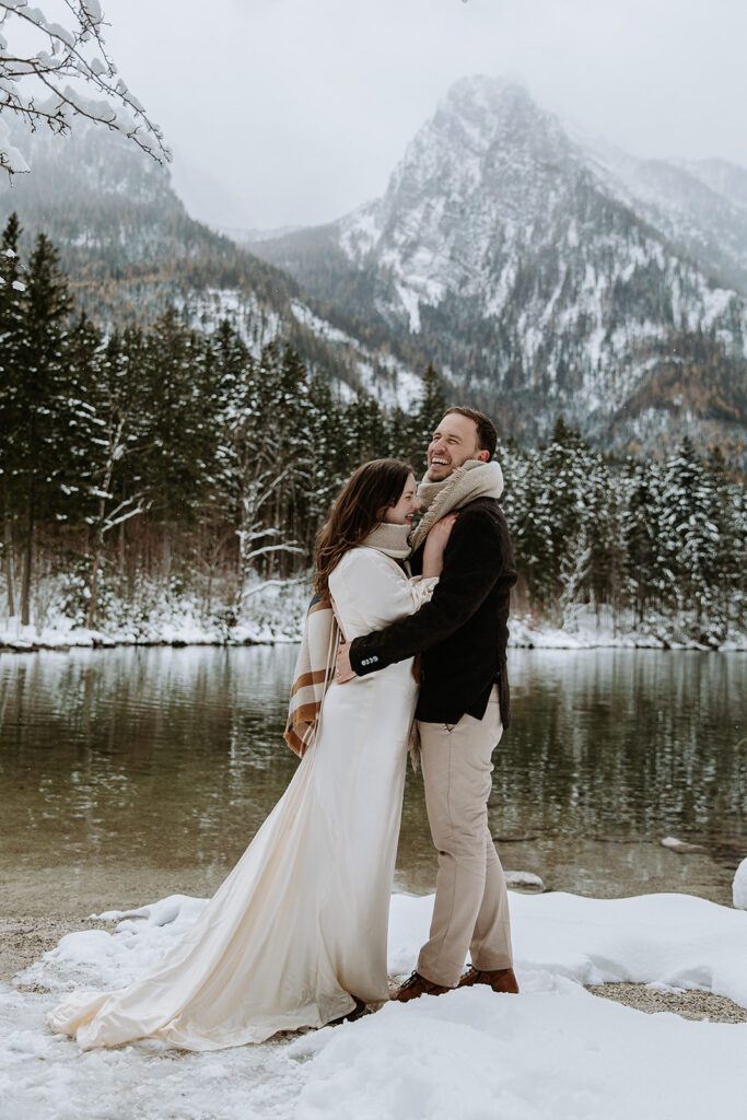 A couple eloping in Berchtesgaden, Germany hug on a snowy winter's day near a snow covered mountain lake