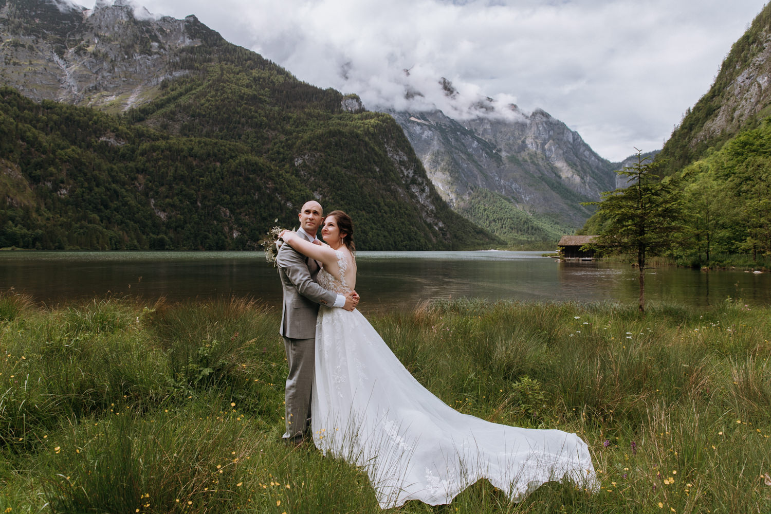 An elopement couple in wedding dress and grey suit stand embracing in a grassy mountain field near a lake in Berchtesgaden, Germany