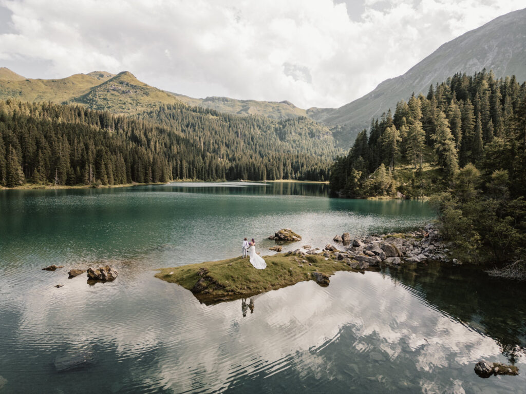 A couple eloping in Austria stand on a small grassy island in the middle of an alpine lake wearing a white wedding dress and white suit