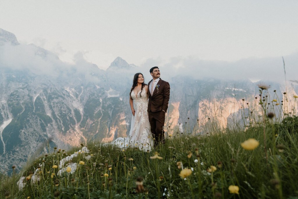 A couple stand looking over a dramatic mountain landscape surrounded by yellow and white wildflowers during their summer elopement in Slovenia