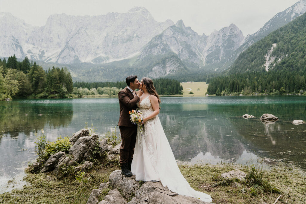 A couple in wedding dress and suit stands kissing on a boulder next to a bright blue lake during their Slovenia elopement