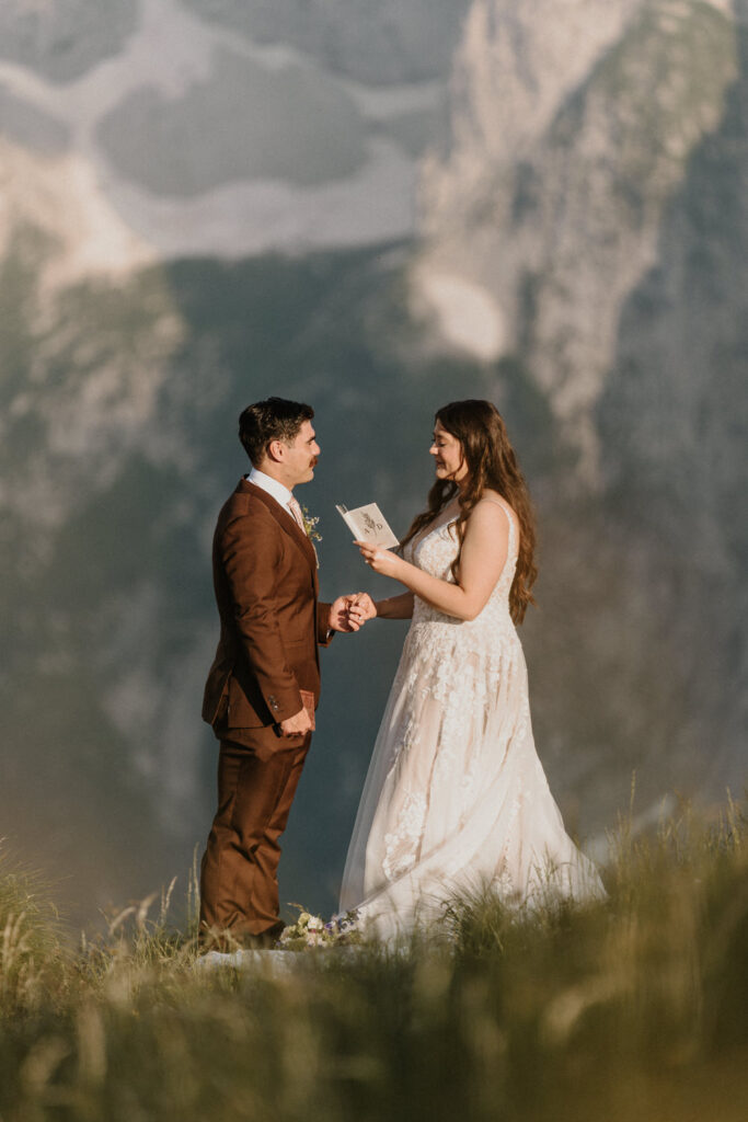 A couple stands holding hands and reading from a white vow book on the side of a grassy mountain during their Slovenia elopement