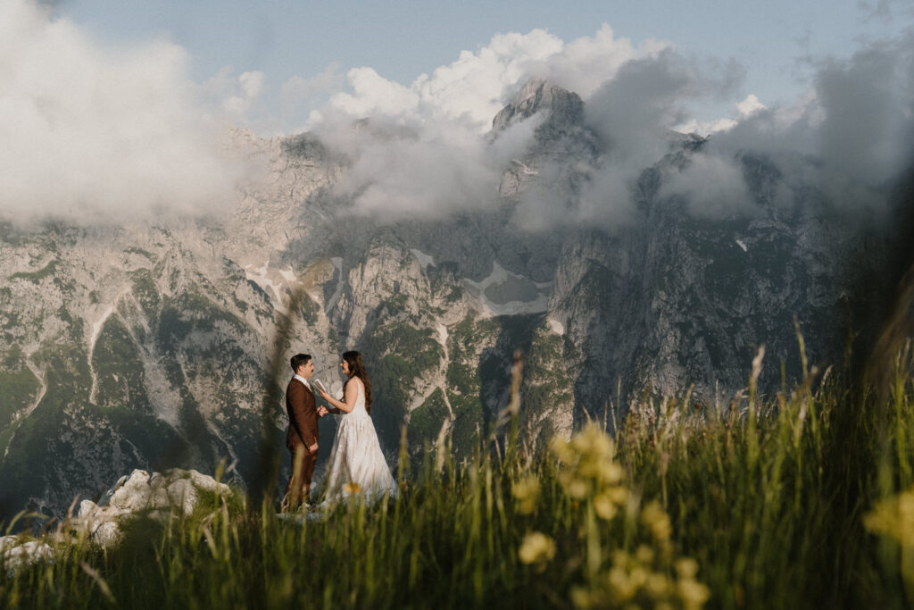 A couple stands holding hands and reading from a white vow book on the side of a green mountain during their Slovenia elopement