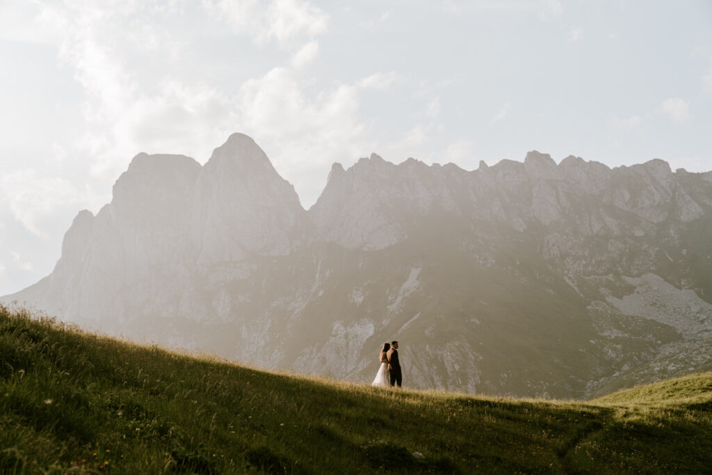 A couple stand small in the middle of the frame on a grassy ridge, surrounded by tall mountain summits and a setting sun on their elopement day