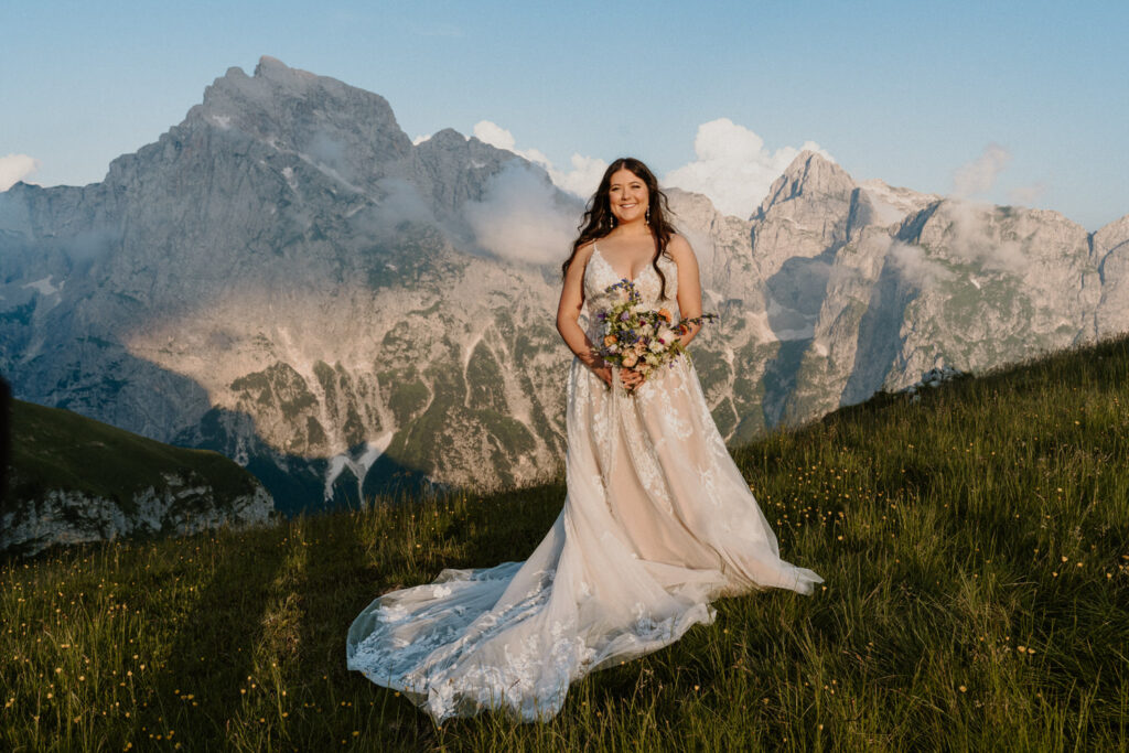 A bride in a wedding dress stands smiling, holding a colorful bouquet on the side of a grassy mountain during her Slovenia elopement