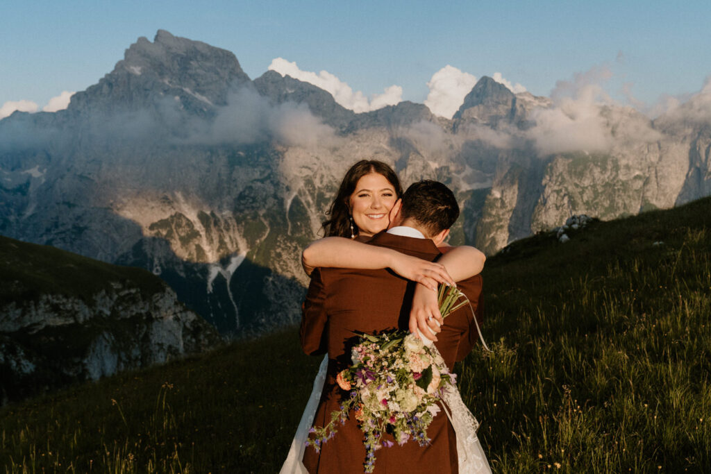 A woman in a wedding dress drapes her arms around her partner and smiles at the camera, with dramatic mountains behind her, during the couple's elopement in Slovenia