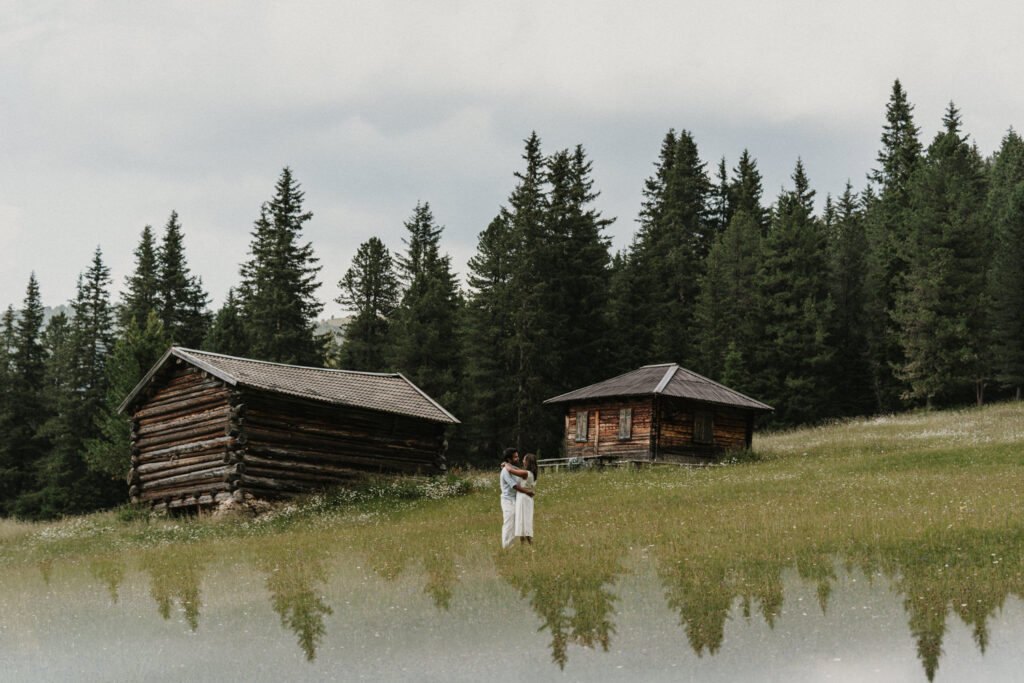 A couple embrace in a grassy field in the Dolomites between two rustic wooden cabins after a surprise proposal.