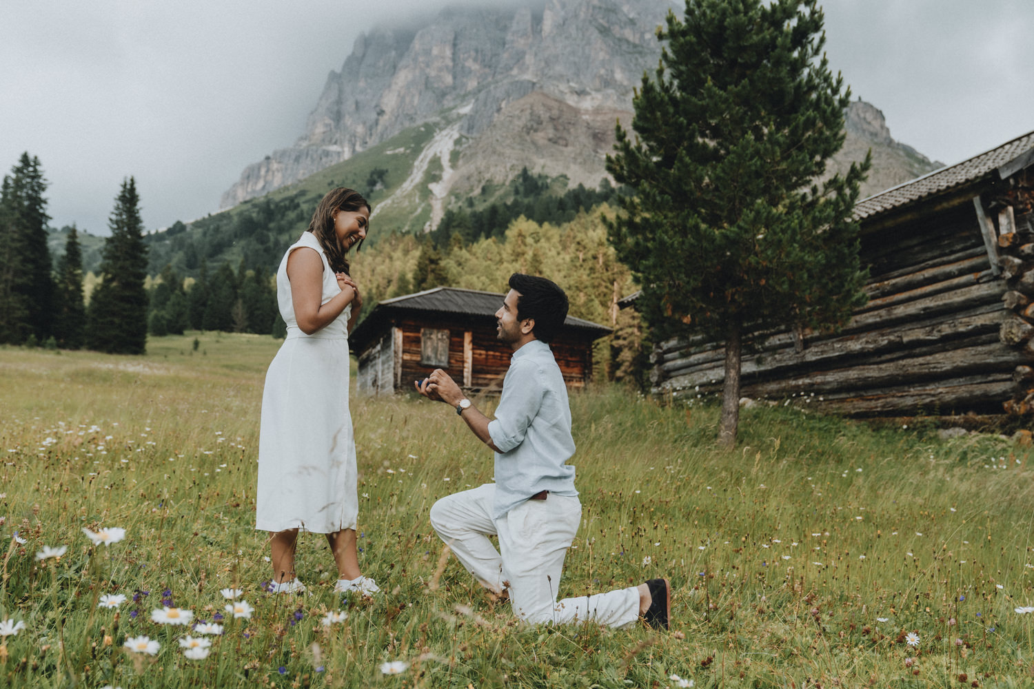 A man in a light blue shirt is on one knee for a surprise proposal to his partner in the Dolomites with mountains and rustic cabins in the background