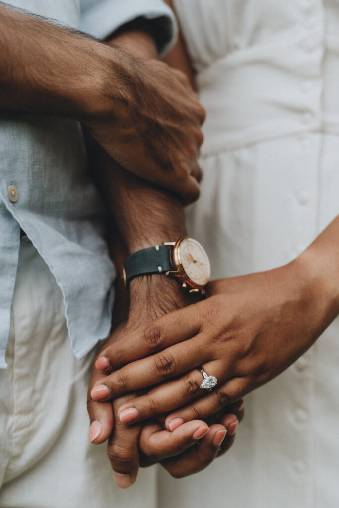 A close up of a darker skinned couple holding hands after their proposal, a large diamond ring visible on her finger