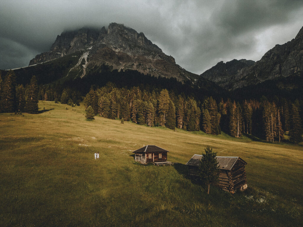 A couple in white outfits stand tiny in the frame, walking in a large grassy field in the Dolomites next to two wooden cabins and tall mountains in the background