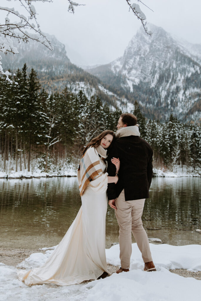 A couple eloping in Austria stand cuddling together on a wintry day near a snow covered lake