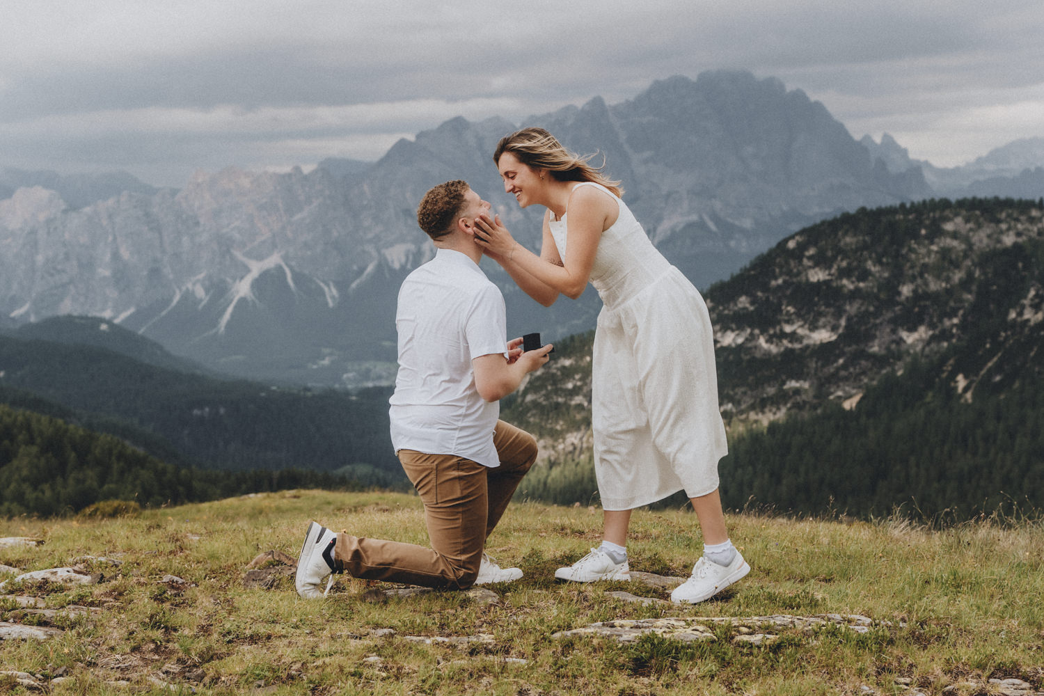 A man in a light blue shirt is on one knee for a surprise proposal to his partner in the Dolomites with mountains and lush fields behind them