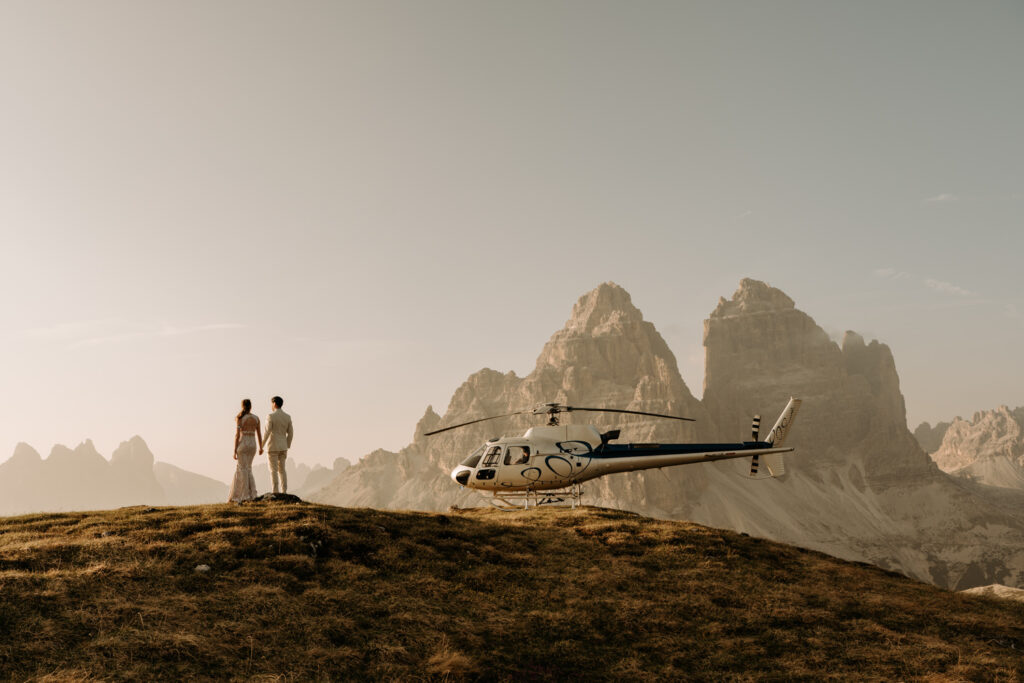 A couple holds hands far away during their elopement facing the Tre Cime in the Dolomites, a helicopter parked next to them