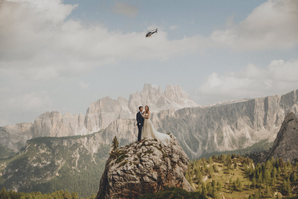 An eloping couple stand on a large boulder in the Dolomites as a helicopter flies over their heads