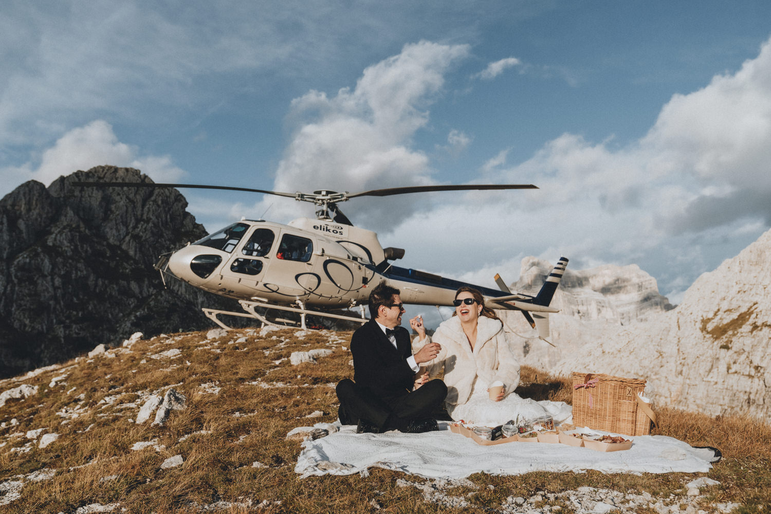 A couple in wedding attire picnic in front of a helicopter on a mountaintop during their Dolomites helicopter elopement day