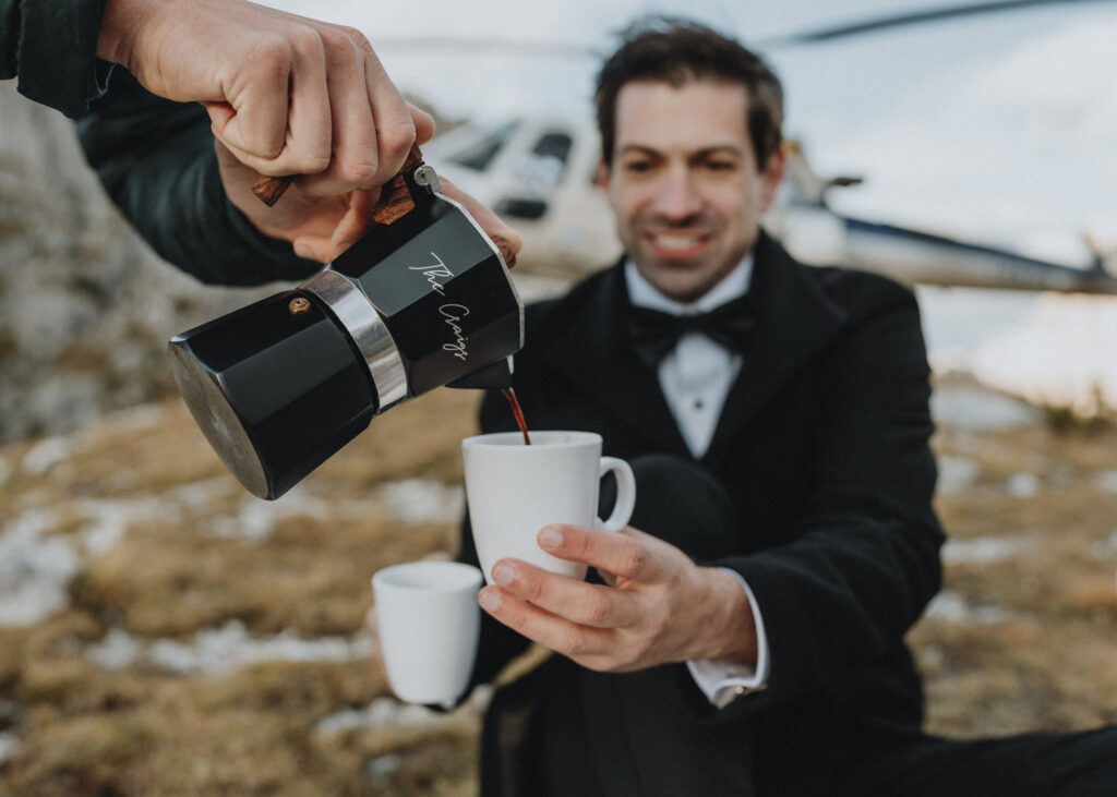 A man in a black tuxedo pours coffee with a helicopter in the background during his helicopter elopement day in the Dolomites