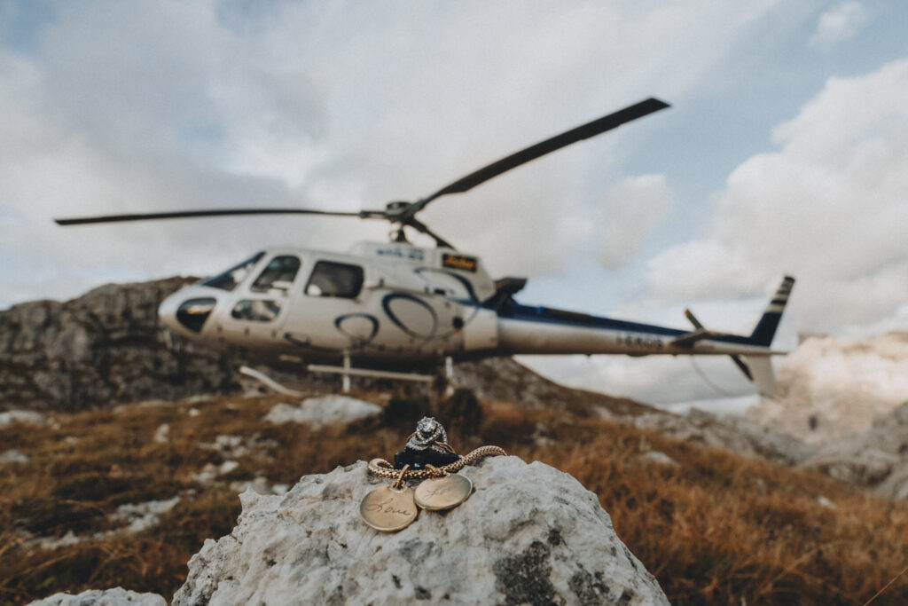 Two wedding rings and other wedding jewelry sit in focus in the foreground with a helicopter rested on the mountain in the background