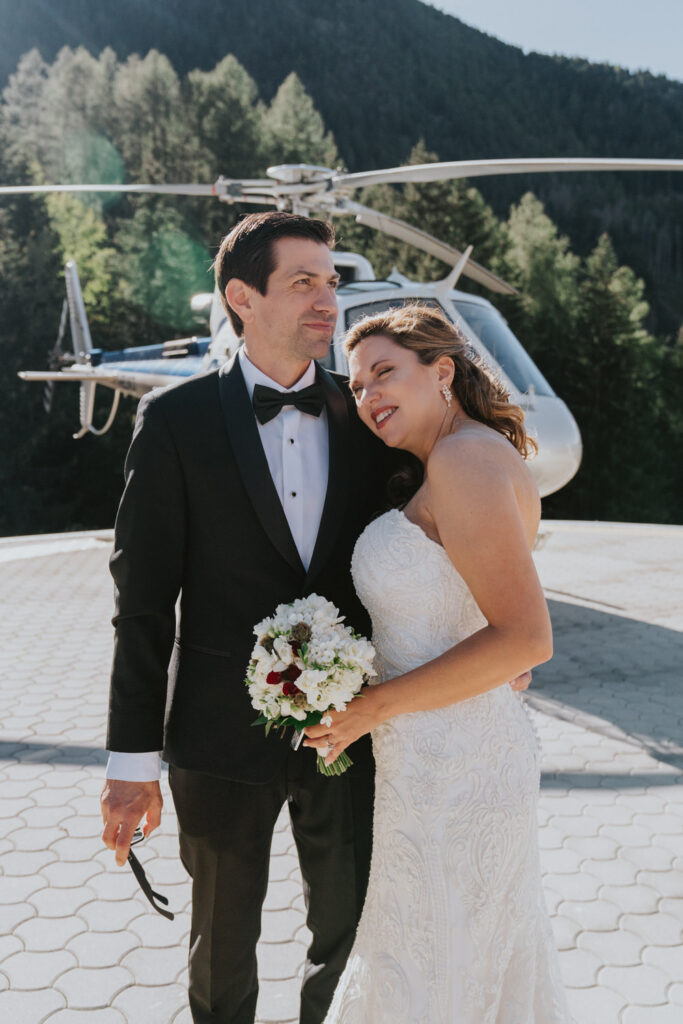 A couple in black tuxedo and white wedding dress embrace outside of a helicopter during their helicopter elopement day