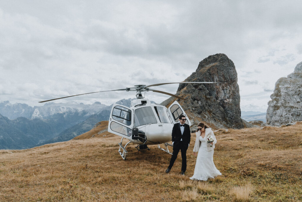 A wedding couple in black tuxedo and white wedding dress walk on a grassy mountain top past a helicopter during their helicopter elopement day