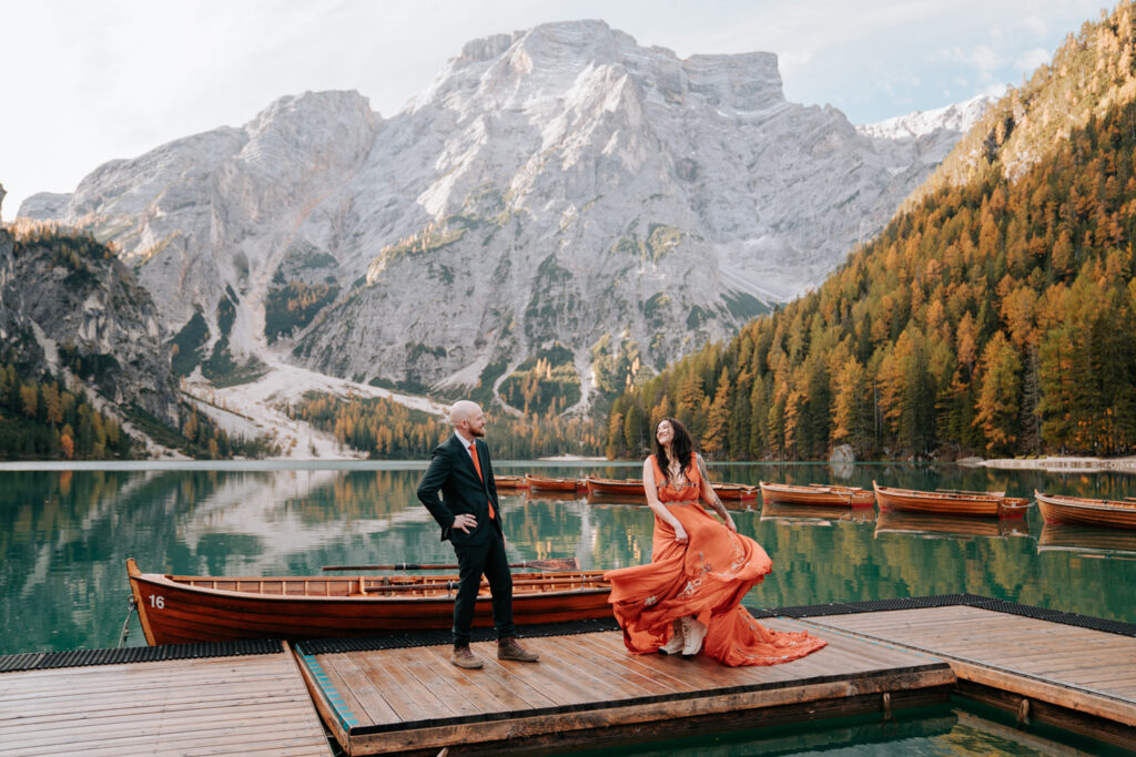 A wedding couple dances in orange dress and green suit at the Lago di Braies boathouse, surrounded by vibrant fall foliage and green water.