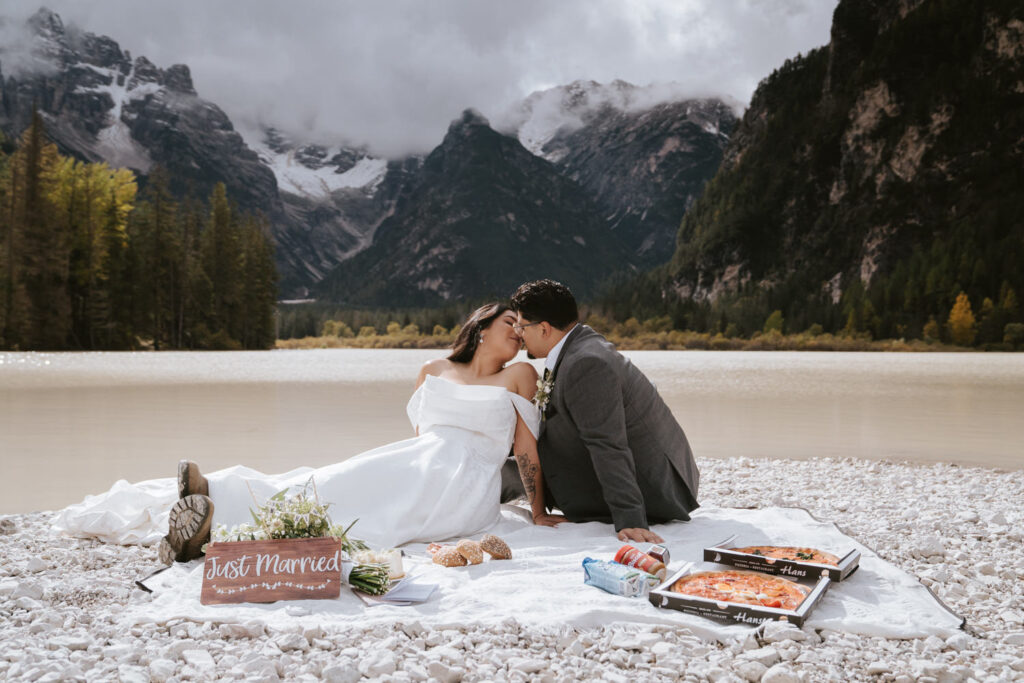 A couple kisses on a picnic blanket on their elopement day in the Dolomites with rich October fall colors on the mountains behind them