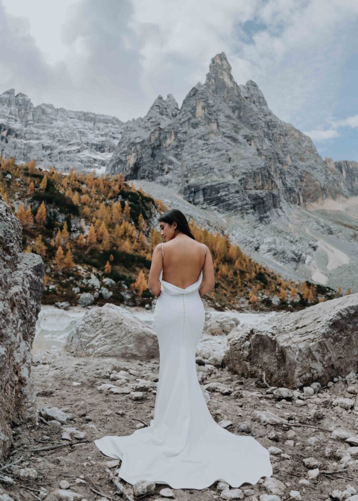 A bride in a white wedding dress poses for her elopement photos in the Italian Dolomites. There are dramatic October fall colors in the trees behind her.