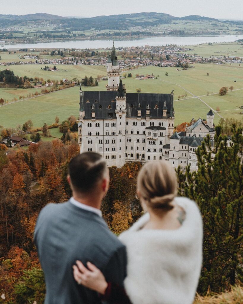 A couple in wedding clothes stands with their backs to the camera looking out over a green landscape with Neuschwanstein castle in the distance