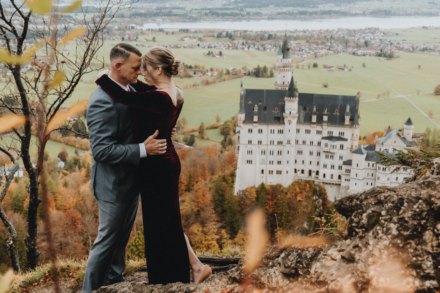 A wedding couple in red velvet dress and grey suit stand face to face on a rocky hilltop with Neuschwanstein castle in the background.