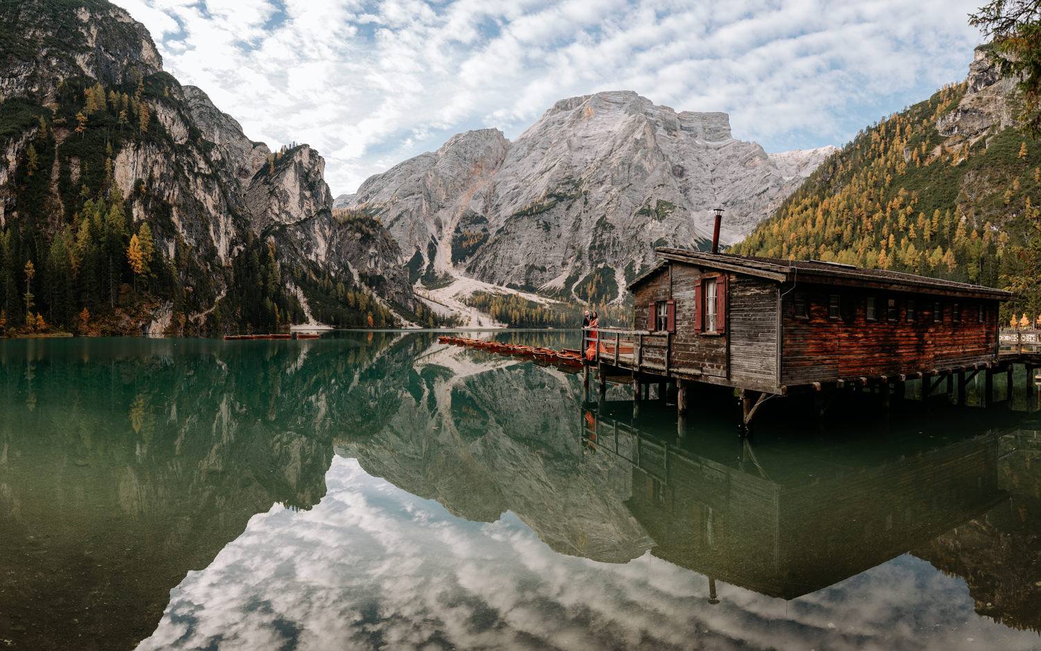 A wedding couple stands on the dock of the wooden boathouse at Lago di Braies, framed by surreal reflections and vibrant fall colors.