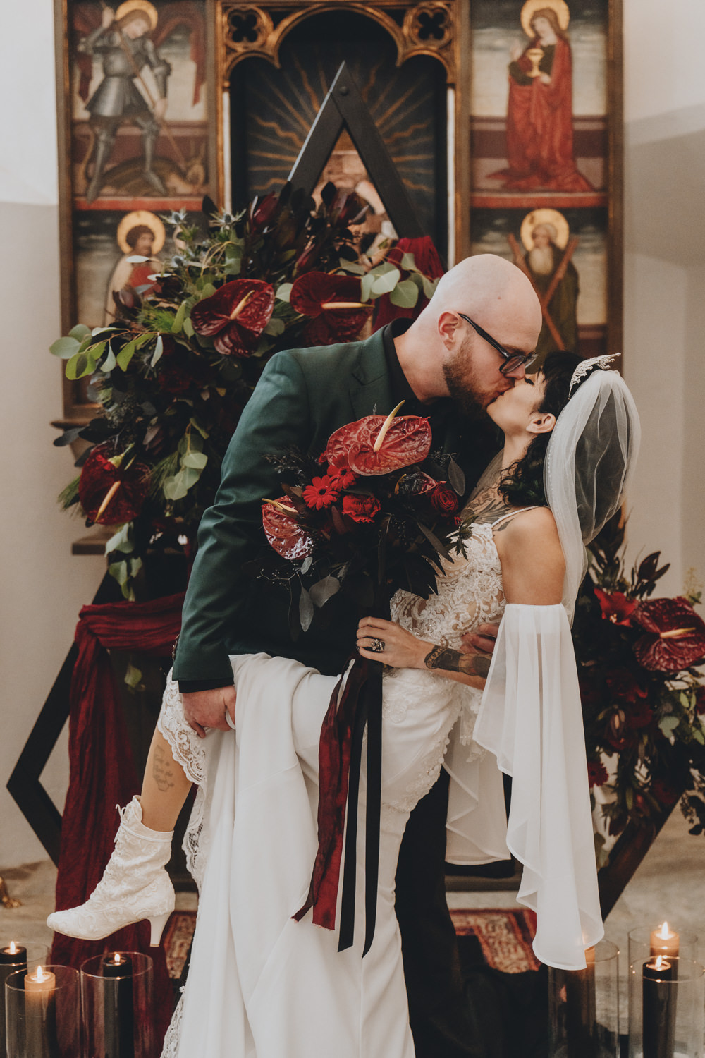 A couple poses in a gothic chapel during their elopement ceremony, surrounded by red and black flowers, lit candles and gothic paintings.