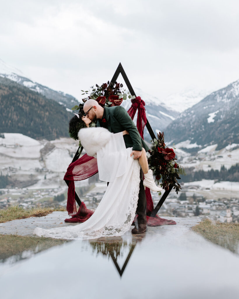 A couple poses in a deep kiss in front of a gothic black wedding arch, snow capped mountains and red flowers during their Austria elopement day