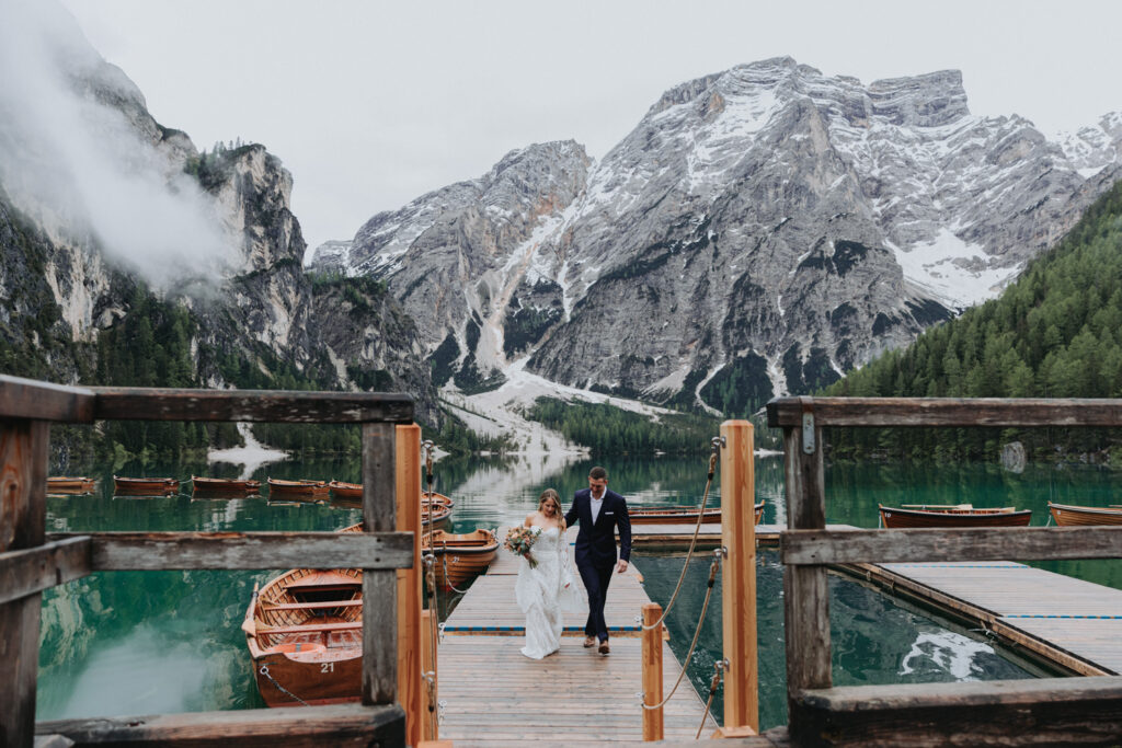 A couple in wedding attire walk along a dock on a lake in the Dolomites after their elopement vows