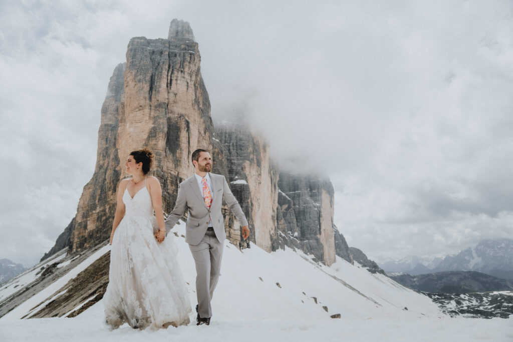 A couple in wedding attire walk away from three dramatic mountain peaks in the Dolomites during their elopement day