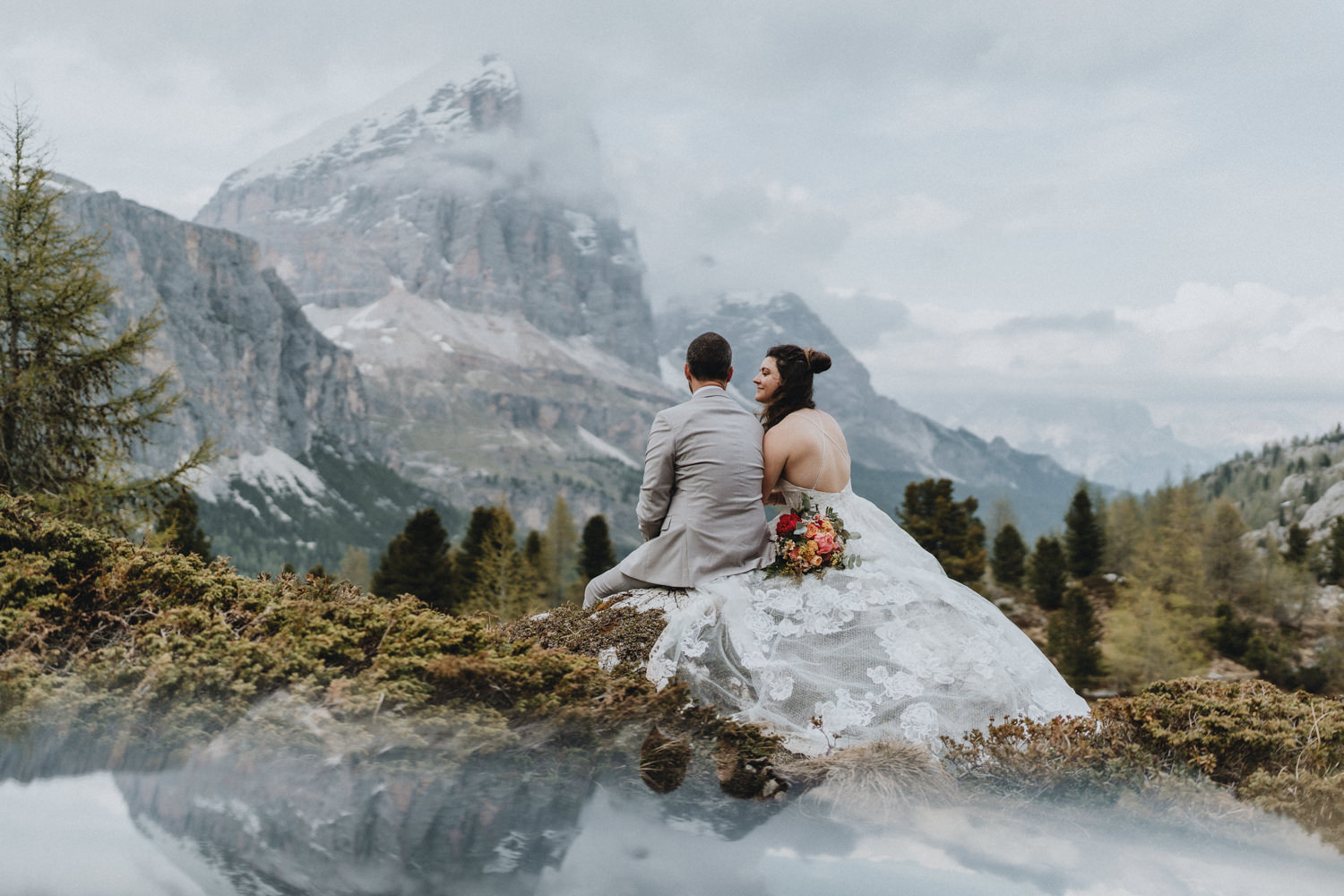 A couple in wedding attire sit on a boulder with dramatic snow covered peaks in the Dolomites in the background