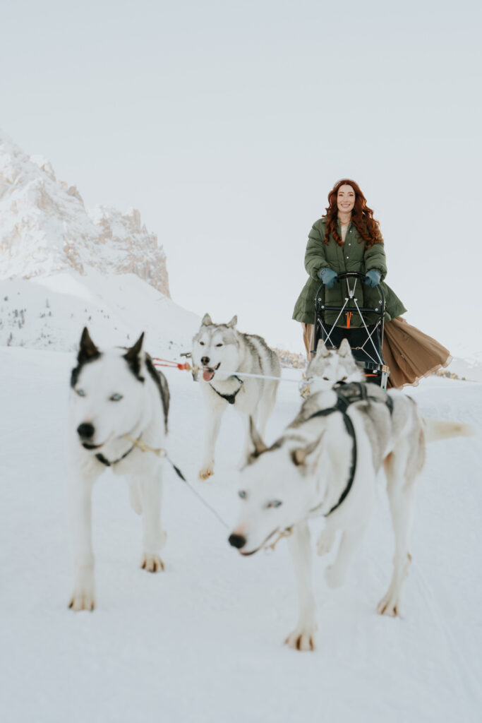 A bride leads a team of husky sleigh dogs through a snowy mountain track during her dog sledding elopement day