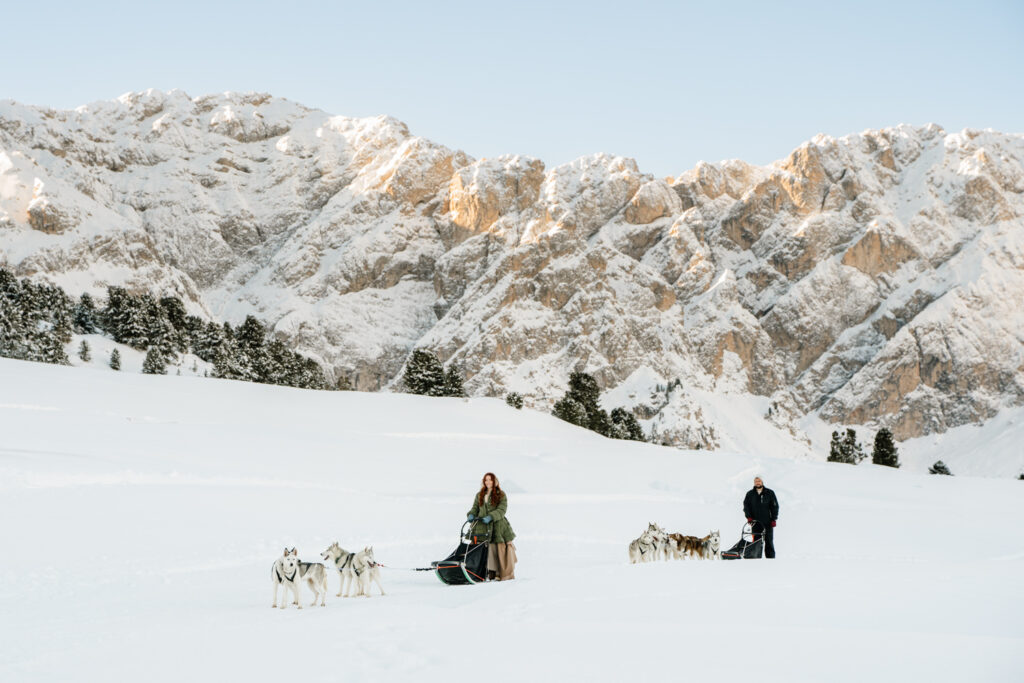 A couple lead two teams of husky dogs across a snowy Dolomites landscape on their dog sledding elopement day