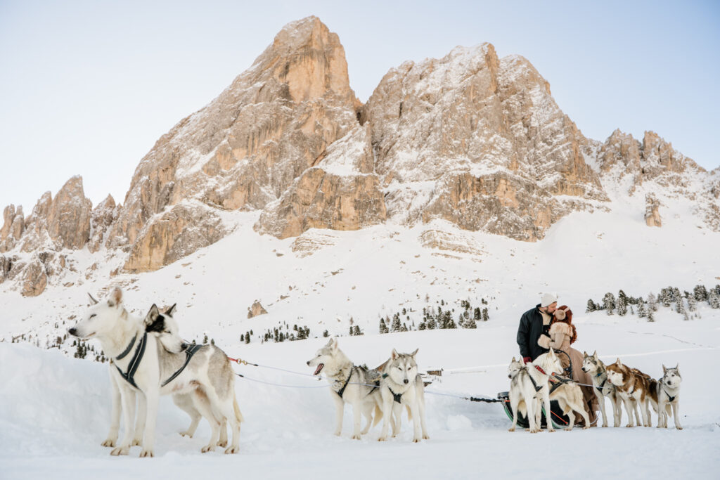 A couple kiss in front of a team of husky dogs with dramatic snow covered mountains behind them on their dog sledding elopement day in the Dolomites