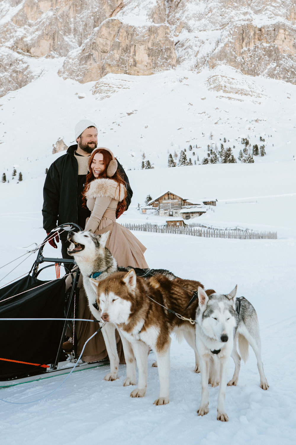 A couple embrace on a dog sled with three husky dogs in front of them and a snowy mountain landscape behind them on their dog sledding elopement day