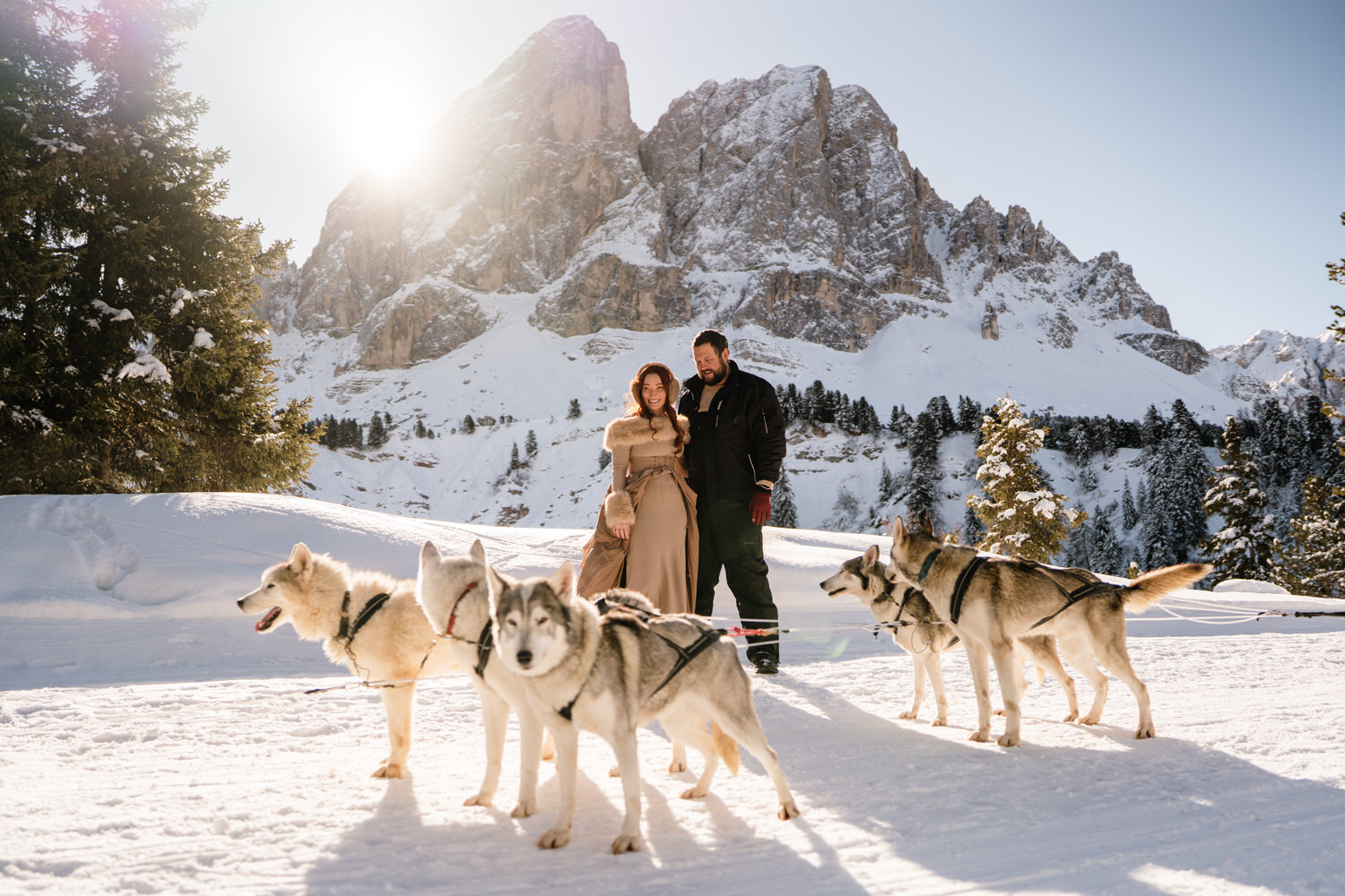 A couple stand in a wintery Dolomites landscape, surrounded by huskies during their dog sledding elopement