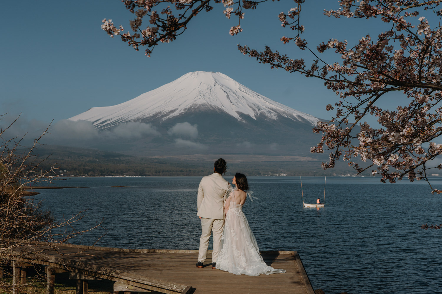 A couple in white wedding clothes stands near the shores of Lake Fujikawago for their Japan elopement, framed by pink sakura and a snowcapped Mt. Fuji in the distance