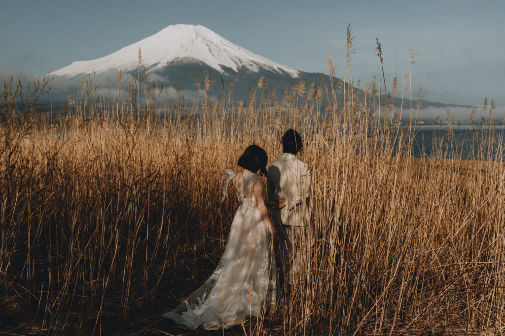 A couple in white wedding dress and suite walk through tall golden grasses toward a snow capped Mt. Fuji during their Japan elopement